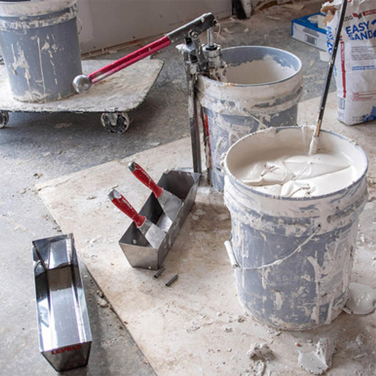 Level 5 joint knives in a mud pan, with a Level 5 mud pump and mud mixer in buckets of drywall compound on a jobsite floor