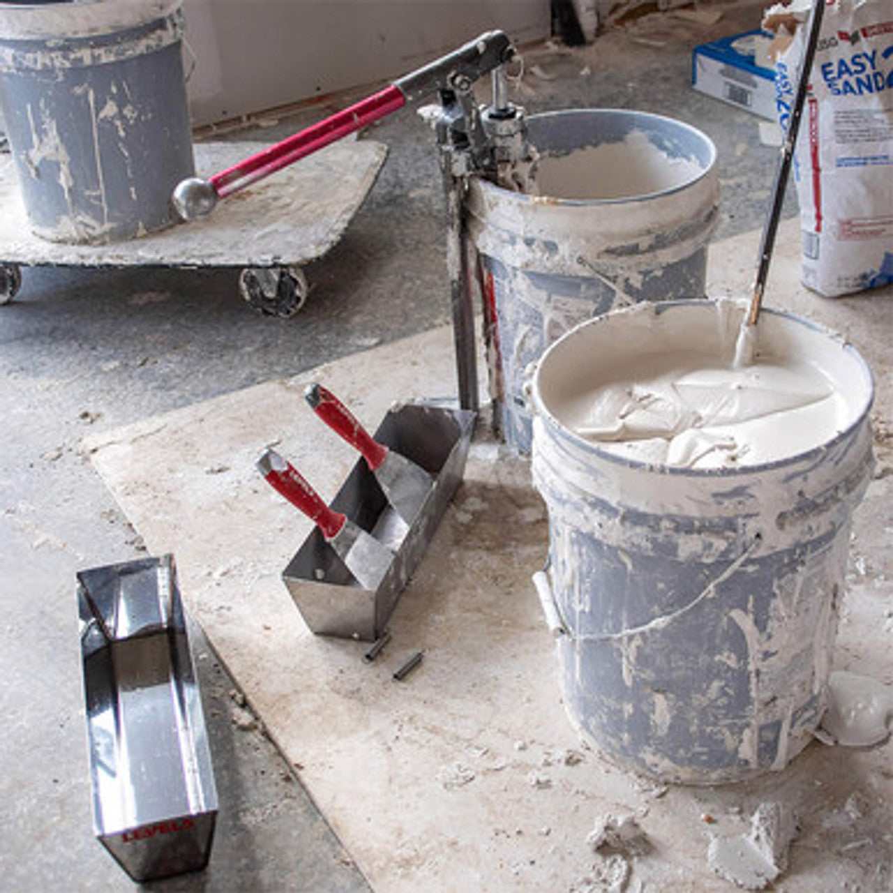 Level 5 joint knives in a mud pan, with a Level 5 mud pump and mud mixer in buckets of drywall compound on a jobsite floor